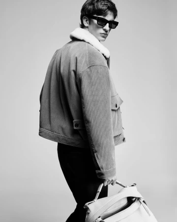 Portrait of a young adult male fashion model in sunglasses, wearing a corduroy jacket with shearling collar and holding a tote bag.