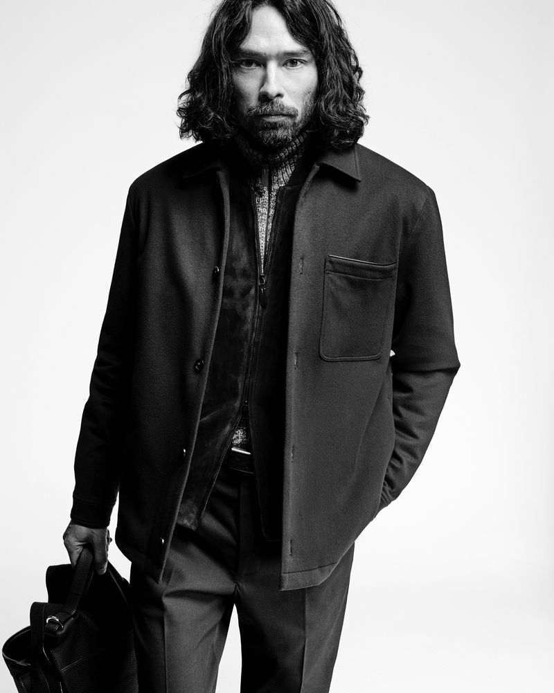 Monochrome portrait of a handsome man with long dark hair, wearing a jacket, against a white background, holding a leather bag.