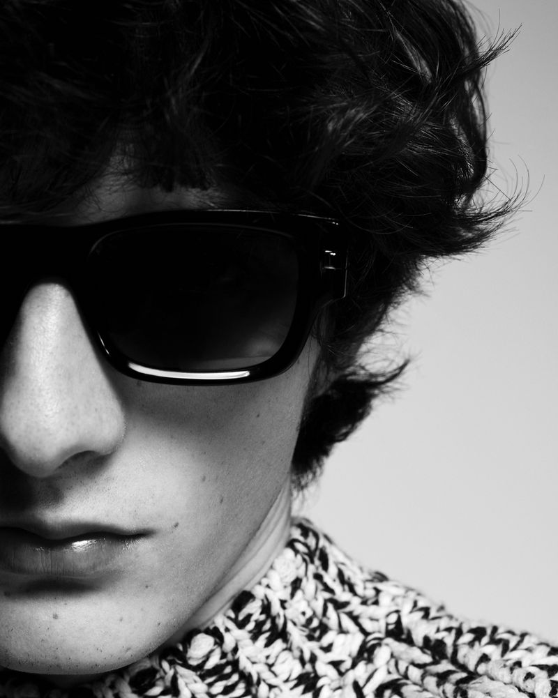 Close-up black and white portrait of a person with dark hair wearing sunglasses and a patterned shirt against a neutral background.