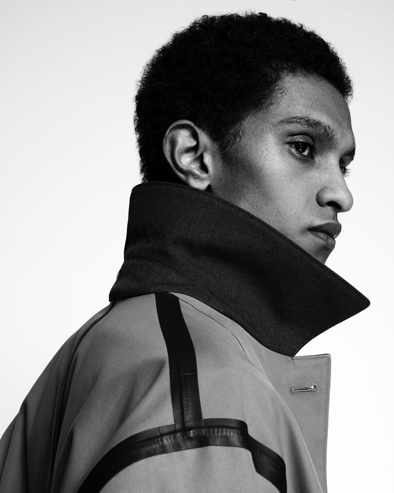 Monochrome portrait of a young adult male model wearing a stylish tan trench coat with bold black stripe accents and a structured collar.