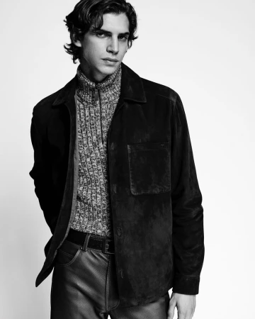 Black and white studio portrait of a young adult male wearing a textured sweater, dark suede shirt-jacket, dark pants, and a belt.