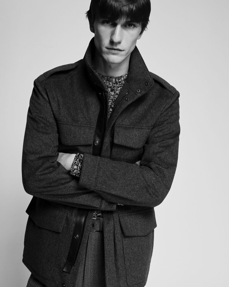 Black and white portrait of a young adult man wearing a wool jacket and patterned sweater in a studio fashion shot with neutral background.