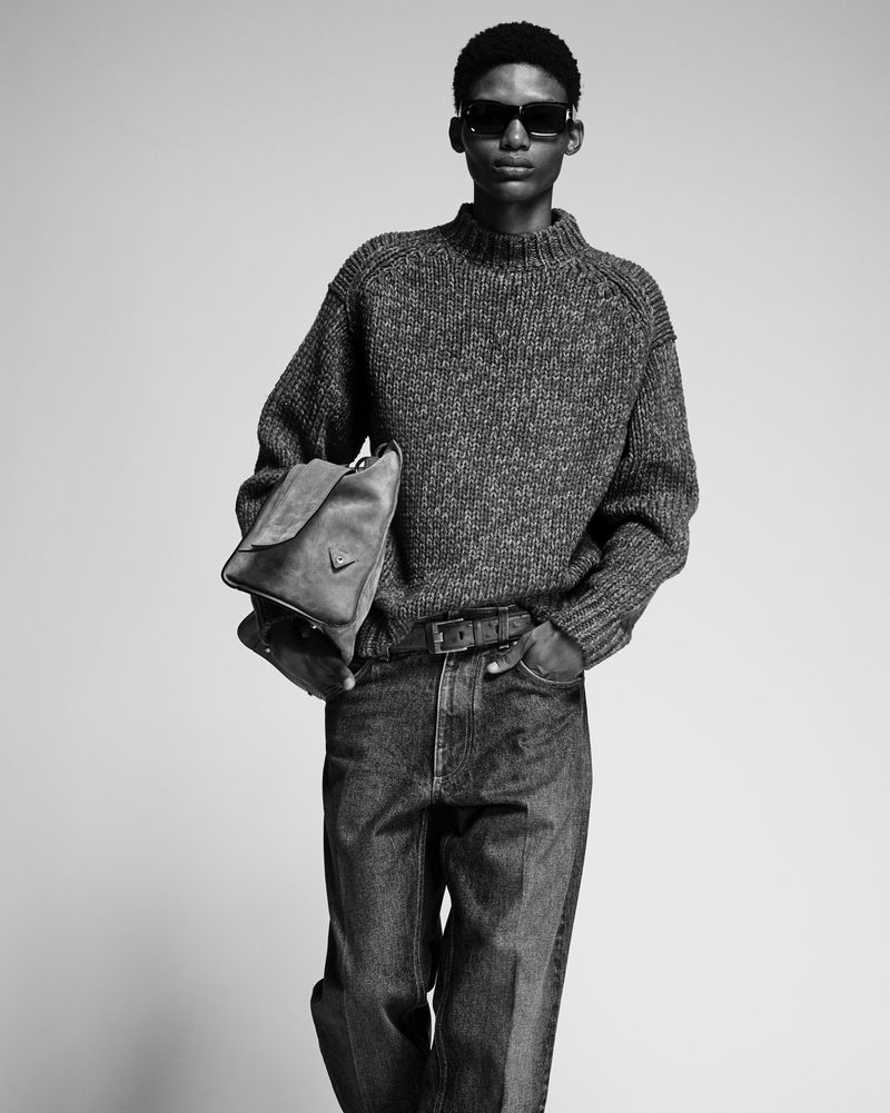 Black and white portrait of a stylish male model in a wool sweater, dark jeans, and sunglasses, holding a leather bag.
