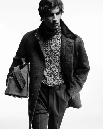 Black and white studio shot of an adult male wearing a wool overcoat and sweater, carrying a duffel bag. Fashion editorial style.