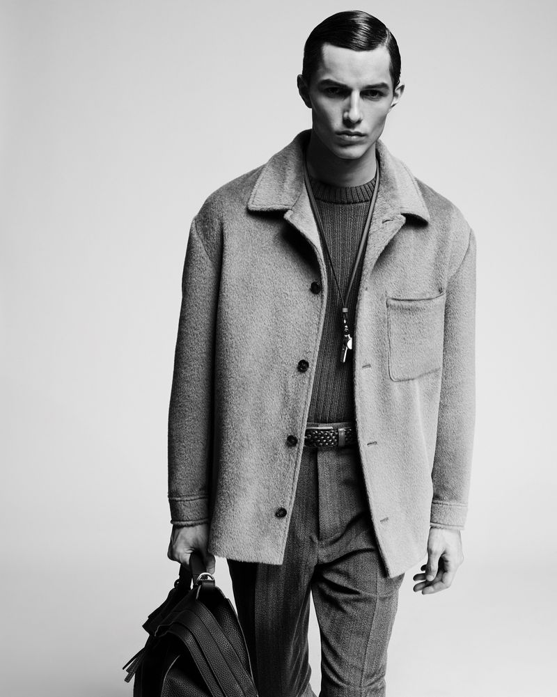 Man wearing a textured grey buttoned jacket and trousers carrying a dark leather bag against a seamless studio background.