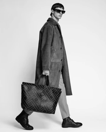 Model in dark sunglasses and long coat carrying a large patterned tote bag against a white seamless background