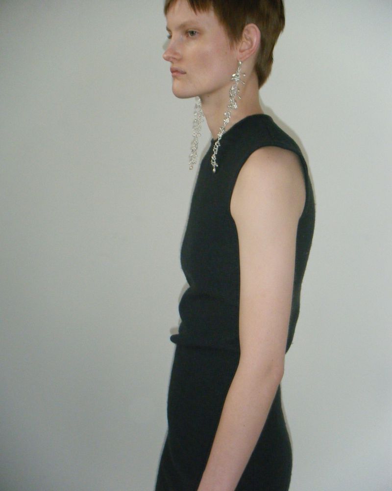 Elegant woman in a black dress and long, dangling earrings, standing in a minimalistic indoor setting with a neutral backdrop.