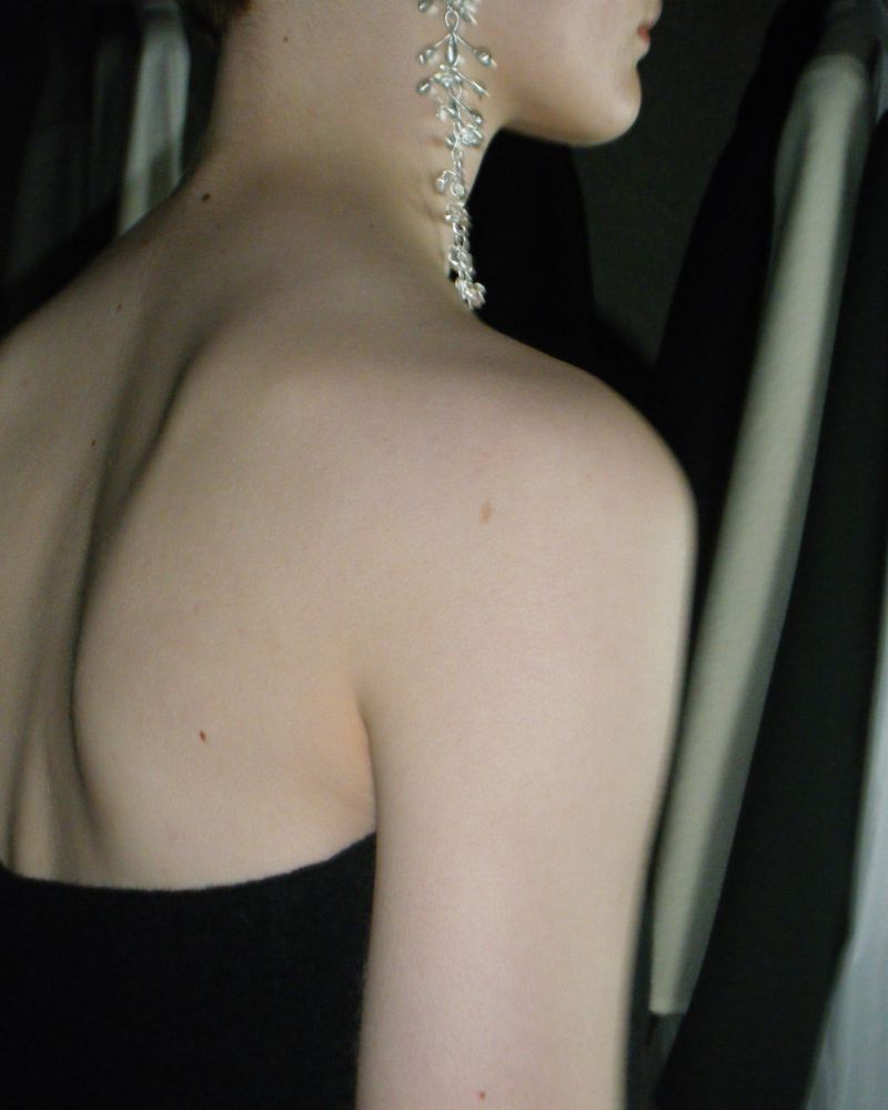 Elegant woman with bare shoulders wearing statement earrings, posing indoors against a background of dark clothing hanging in a closet.