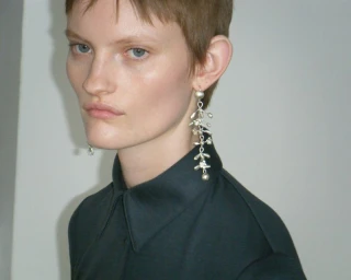 Close-up portrait of a model with short brown hair wearing a dark shirt and long silver drop earrings against a plain background