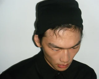 Model wearing a black cap and dark shirt against a white seamless background for Rier Backstage Winter 2025.