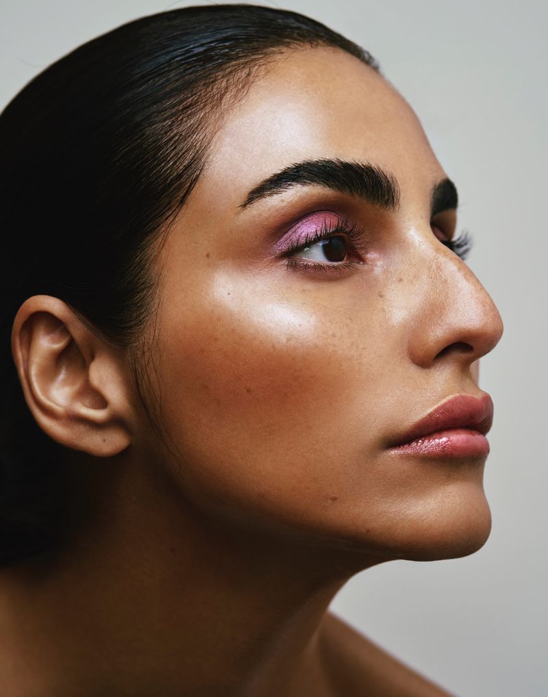 Close-up portrait of a woman with sleek dark hair, wearing pink eye shadow and lipstick, against a light gray background.