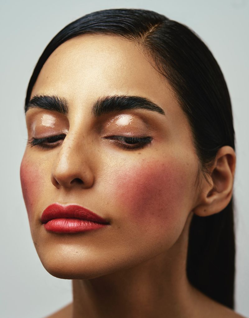 Close-up beauty shot of an adult woman with red lipstick, glossy eyelids, pink blush, and dark hair against a neutral background.