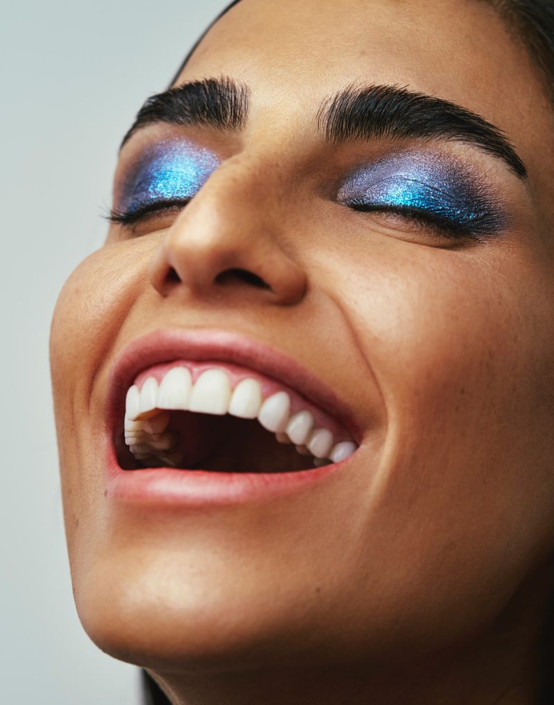Close-up portrait of a woman with vibrant blue glitter eyeshadow and a joyful expression, captured against a clean backdrop.