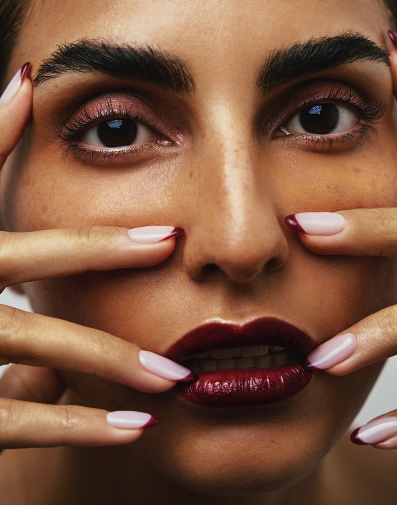 Close-up portrait of a woman with dark red lipstick and manicured fingernails against a neutral backdrop in a beauty style photograph.