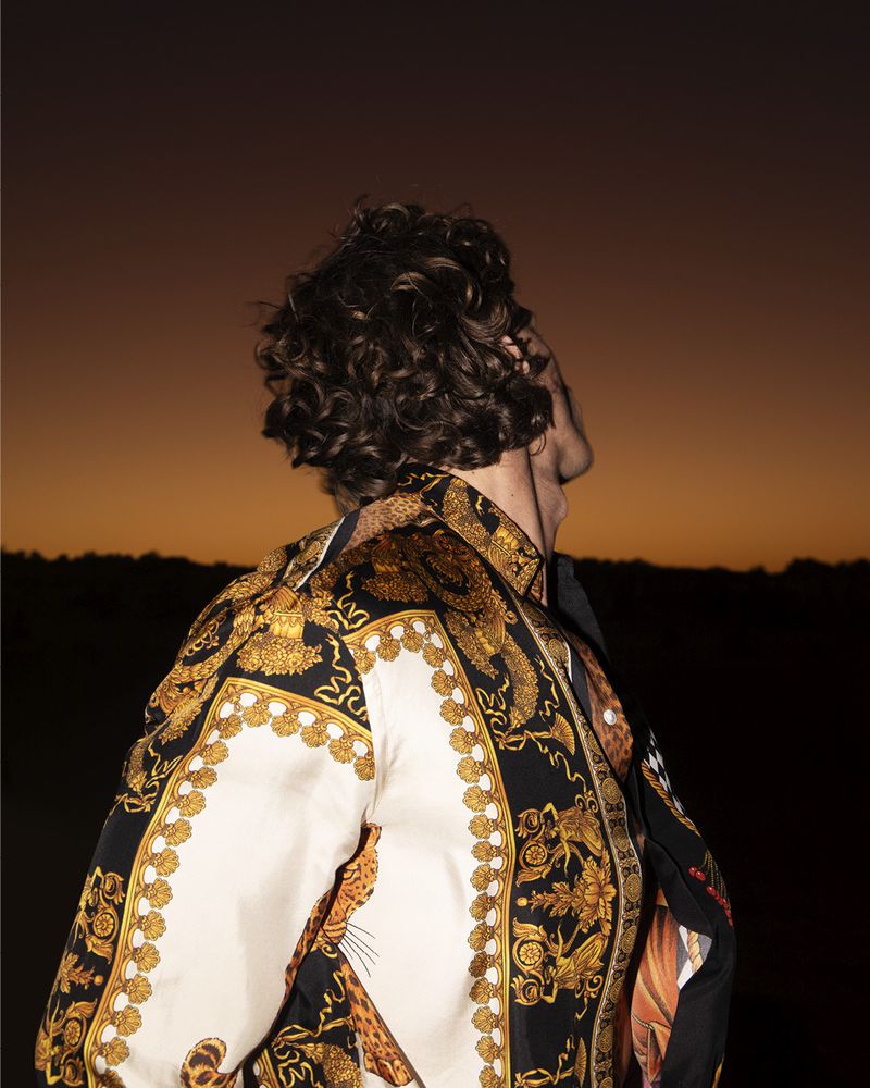 Back view of a stylish adult male with curly hair, wearing a patterned shirt against an outdoor sunset background at twilight.