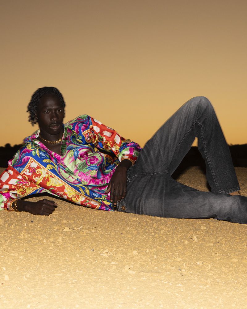 Dark-skinned man lounges on sand in jeans and a colorful patterned shirt, jewelry visible, in an outdoor setting at twilight.