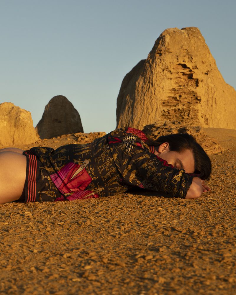 Adult laying on the ground in a desert with rock formations in the background at sunset with a patterned jacket.