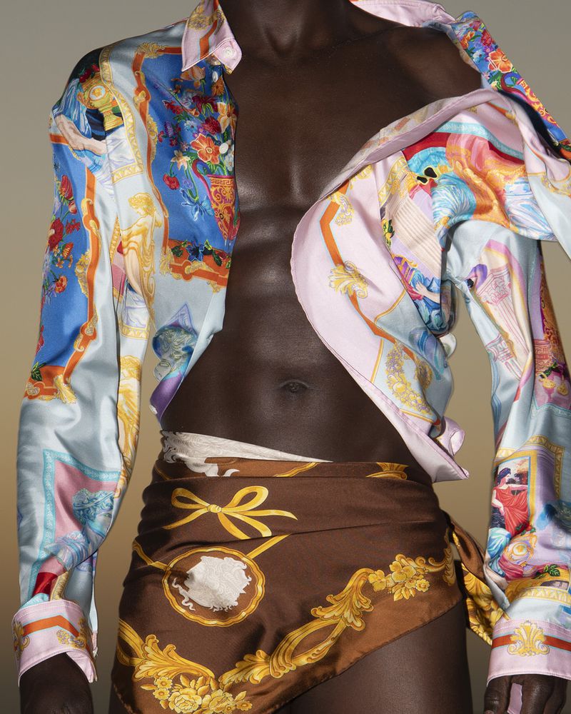 Dark-skinned man with a bare torso wearing a patterned shirt and scarf tied around waist, posing in a studio with diffused lighting.