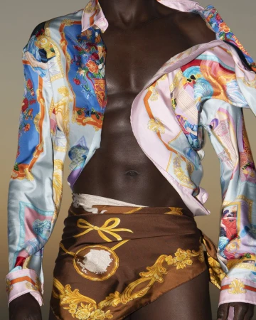 Dark-skinned man with a bare torso wearing a patterned shirt and scarf tied around waist, posing in a studio with diffused lighting.