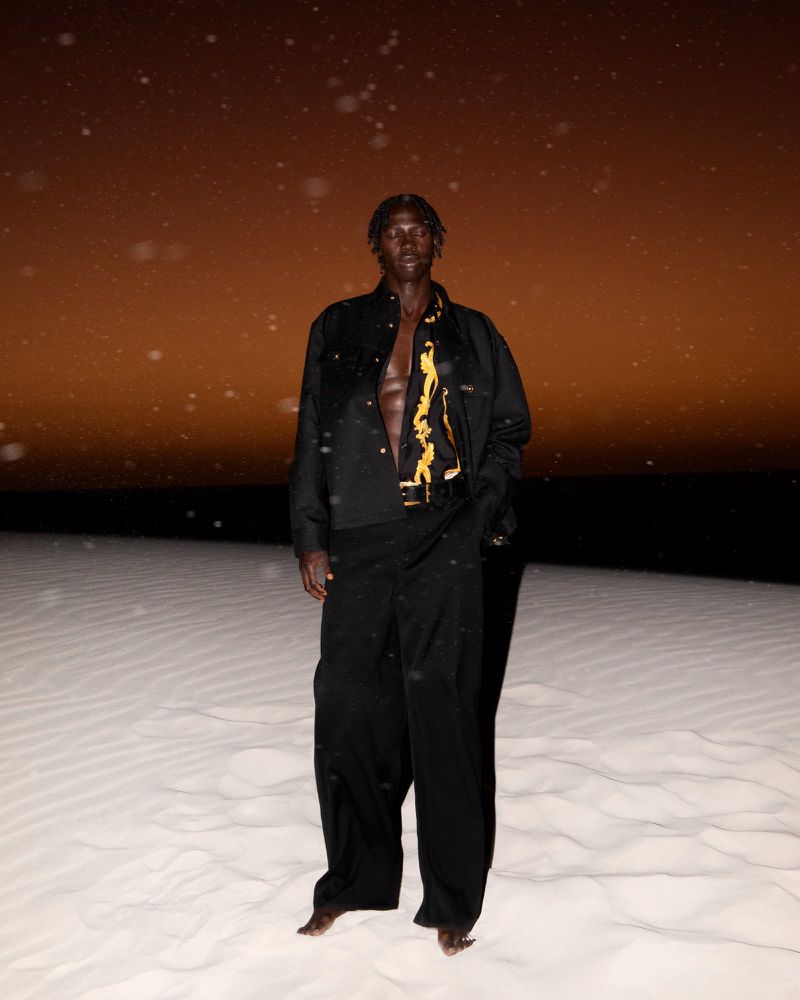 Full-length portrait of a man in black clothing posing in a sandy environment beneath a warm sky, captured in an editorial style.
