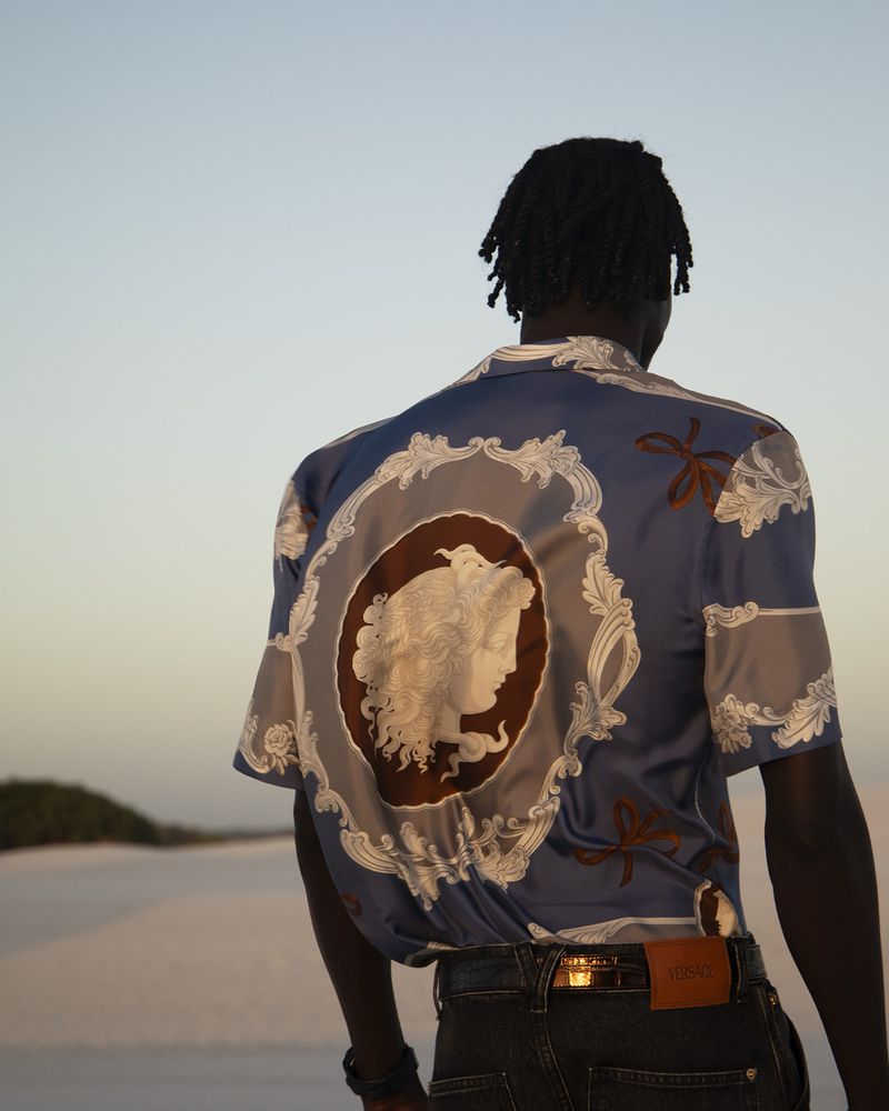 Back view of a Black adult male wearing a patterned shirt with jeans, standing outdoors in a sandy landscape under a blue sky.