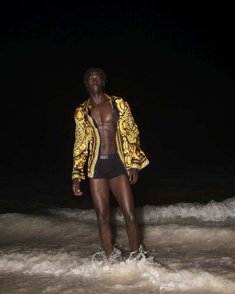 A Black male model stands in ocean waves at night, wearing black briefs and an open, ornate yellow-and-black Versace shirt.