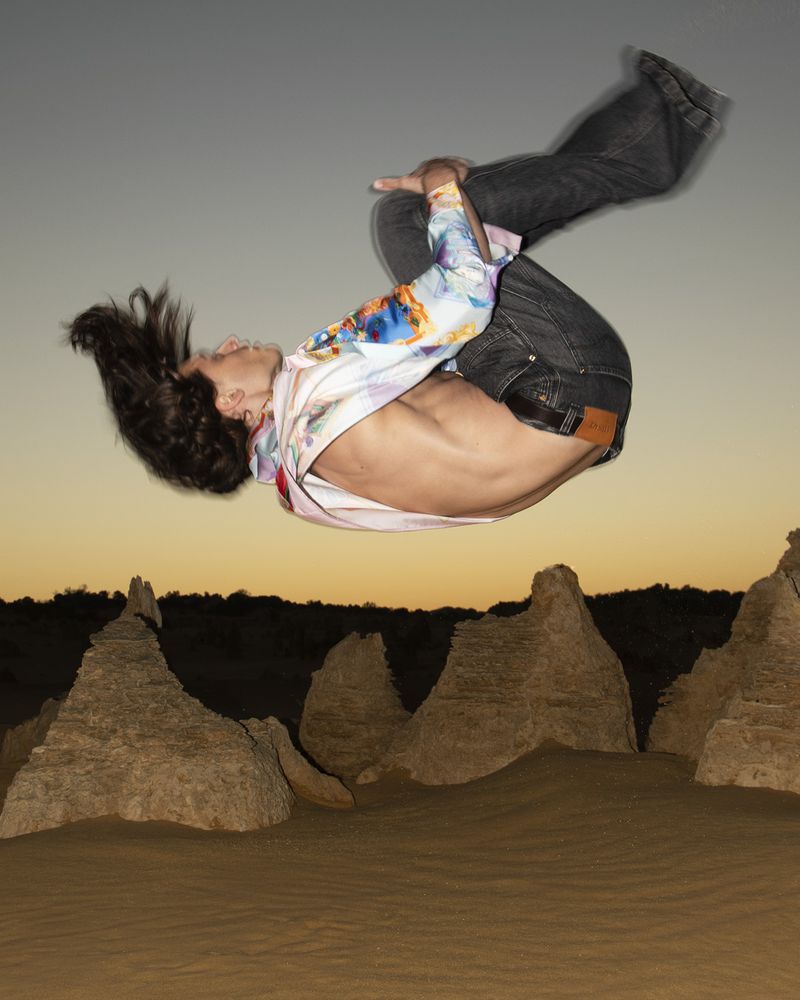 Adult male does backflip over the Pinnacles Desert in Western Australia at dusk; wearing jeans and a patterned long-sleeved shirt.