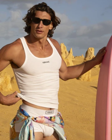 Male fashion model poses on a sandy beach wearing a Versace tank top, briefs, and sunglasses while holding a pink surfboard.