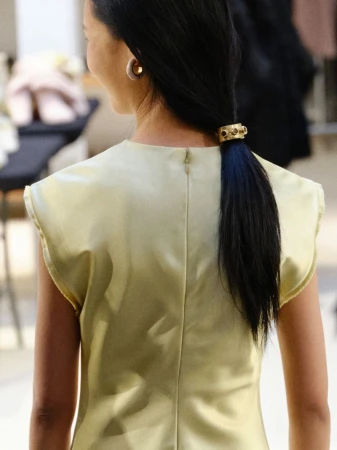 Back view of a woman with black hair styled in a low ponytail with a gold cuff, wearing a sleeveless gold satin top indoors.