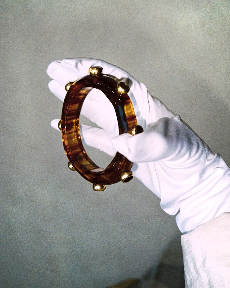 A gloved hand holds a bracelet made of tortoiseshell-patterned material and gold spheres against a grey background in a studio setting.