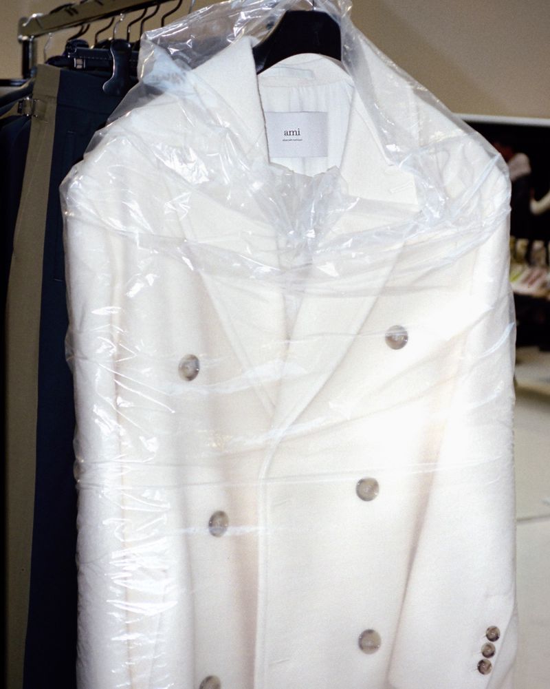 Double-breasted cream-colored overcoat on a hanger in a plastic garment bag on a clothing rack inside; fashion still life.