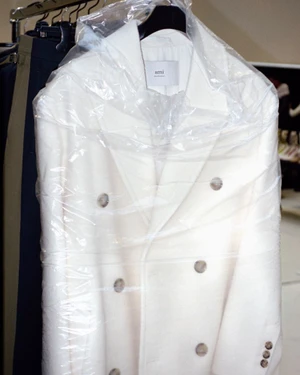 White double-breasted coat in a clear plastic garment bag hanging on a rack for Ami Fall-Winter 25 Fittings.