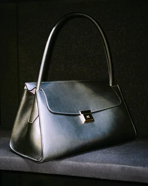 A structured dark leather handbag with a top handle and metallic clasp resting on a dark surface against a dark textured background.