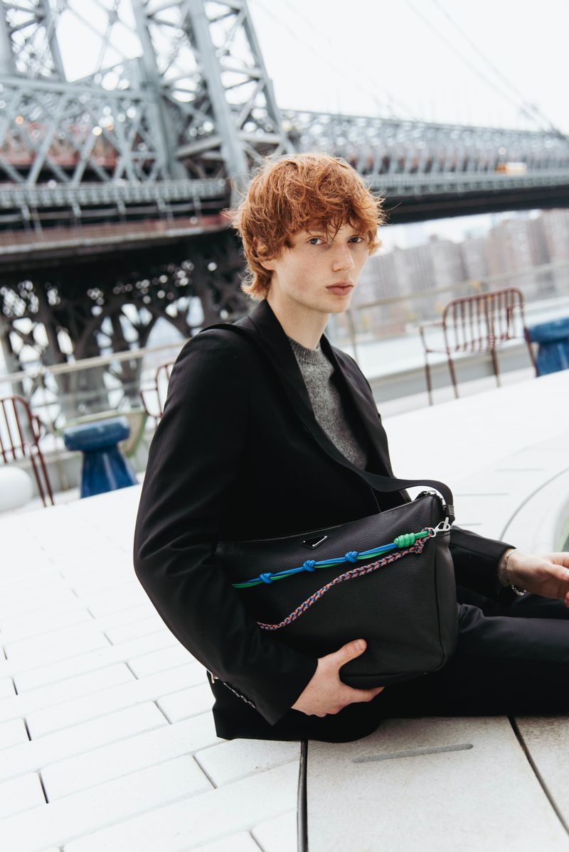 Fashionable man with ginger hair poses with a black leather messenger bag with colourful rope accent, city and bridge in background.