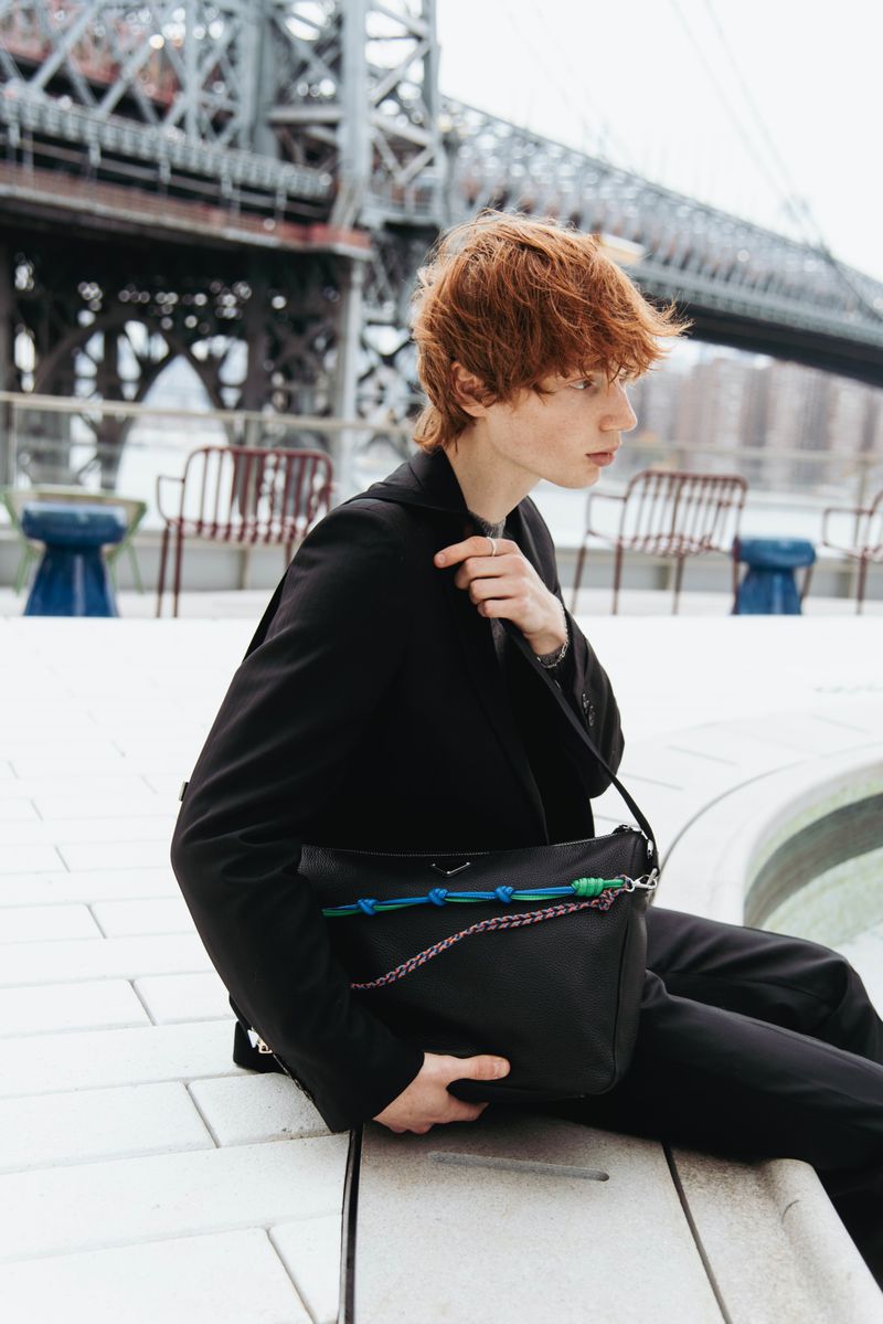 A person with red hair in a black suit sits outside with a black shoulder bag and a bridge in the blurred background.