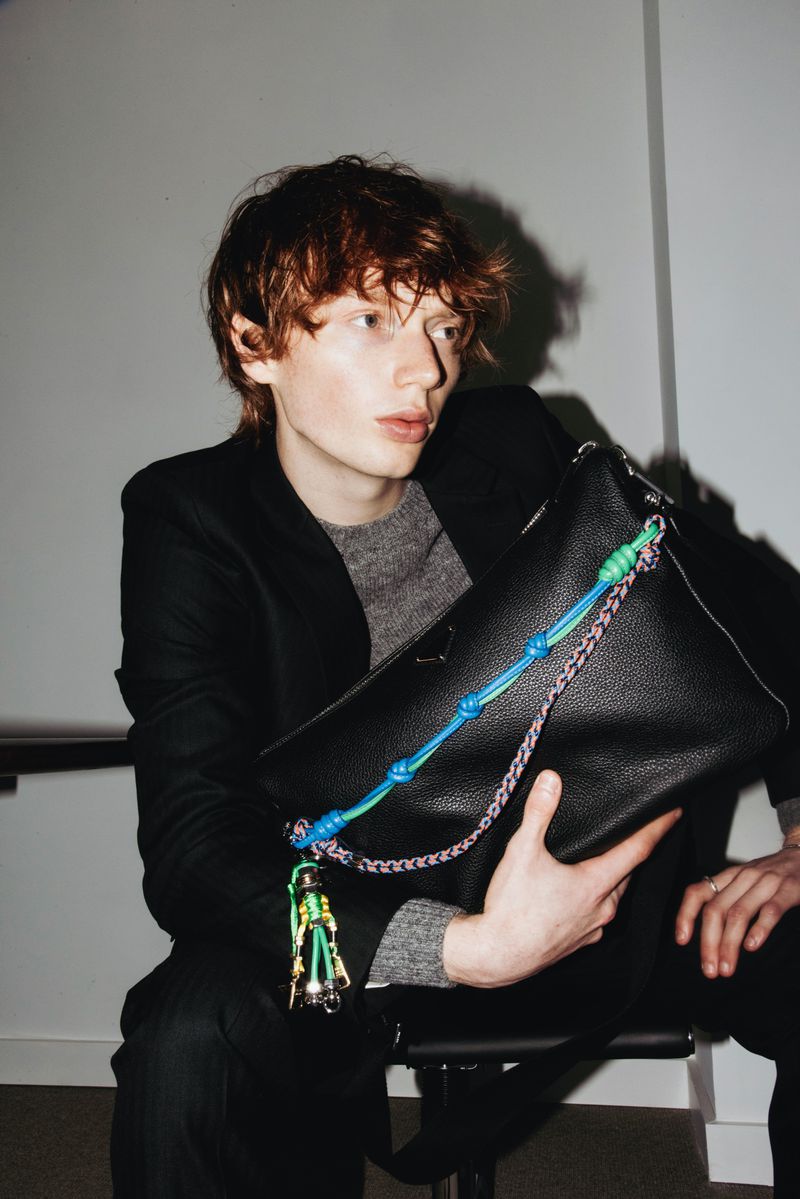 Red-haired male model in a grey sweater and black suit holds a large black leather bag with chain and knotted rope detail in a photo studio.