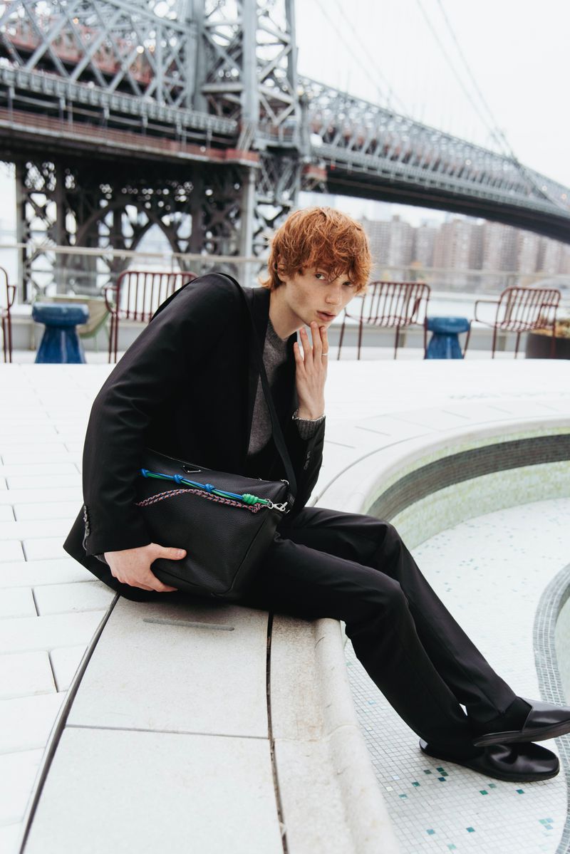 Male fashion model with red hair in dark suit poses with bag in urban setting with bridge visible in the background, looking at camera.