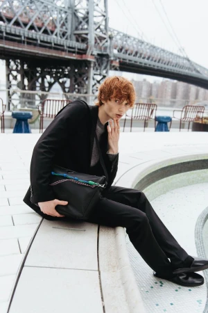 Male fashion model with red hair in dark suit poses with bag in urban setting with bridge visible in the background, looking at camera.