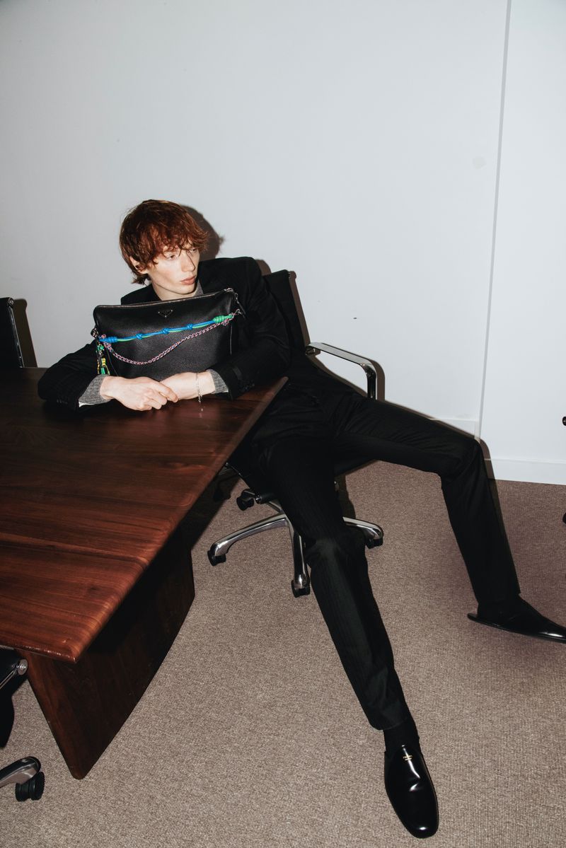 Young adult male in a suit poses with a black handbag on a brown table in a photo studio setting with simple lighting.