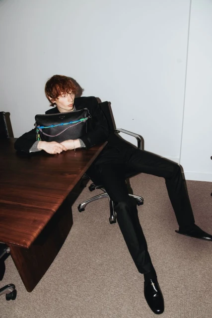 Young adult male in a suit poses with a black handbag on a brown table in a photo studio setting with simple lighting.