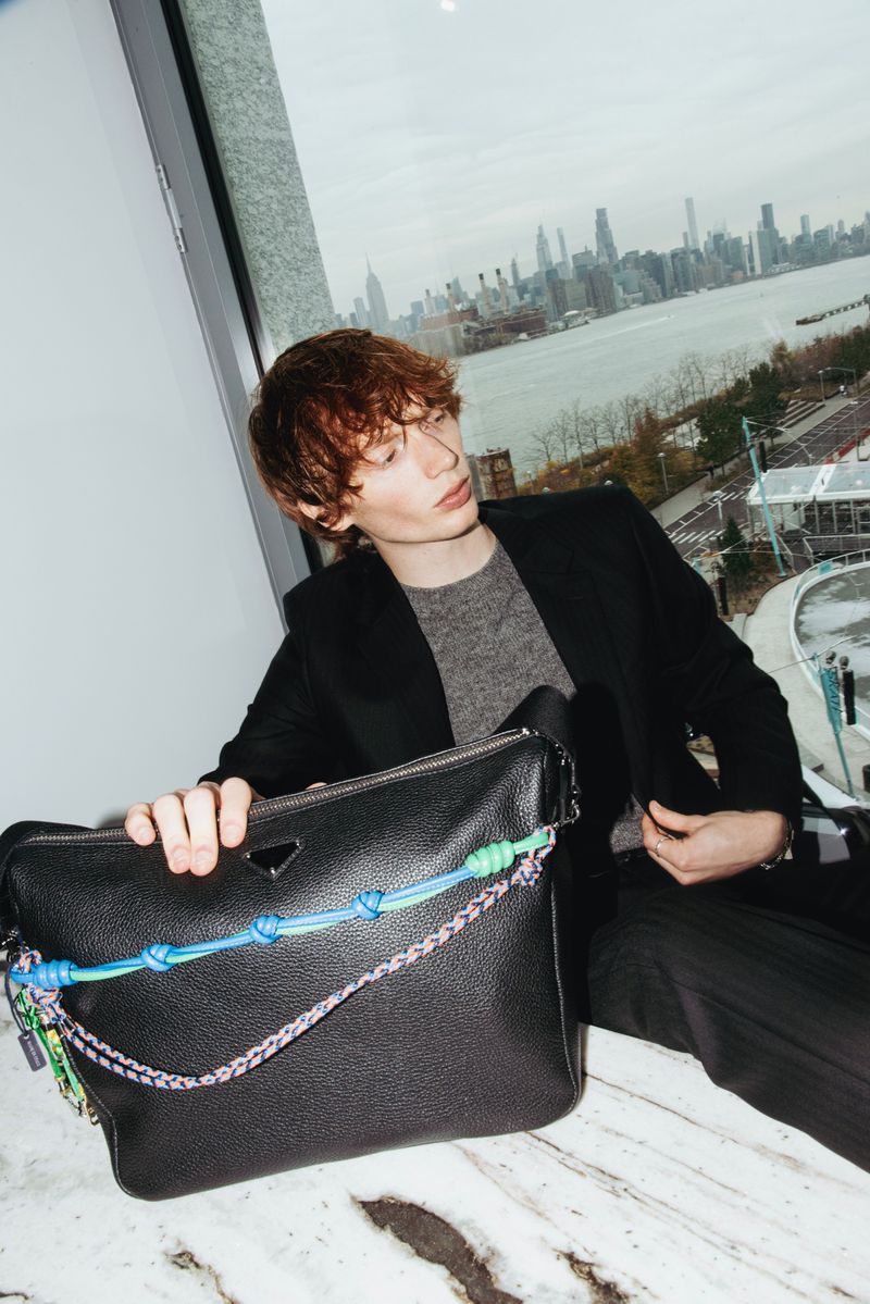 A young man poses indoors with a black leather shoulder bag, city skyline visible through a window providing natural light.