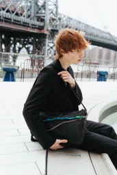 Young man with reddish hair wearing a black jacket holding a black shoulder bag outdoors near a bridge.