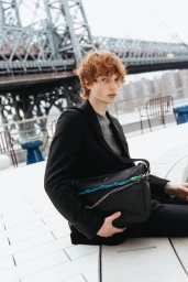 Model Calum Harper wearing a black blazer holding a black leather shoulder bag with a large bridge in the background