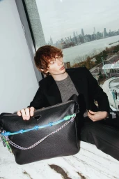 A young man holding a large black leather bag with colorful braided straps, seated indoors near a window overlooking a city skyline.