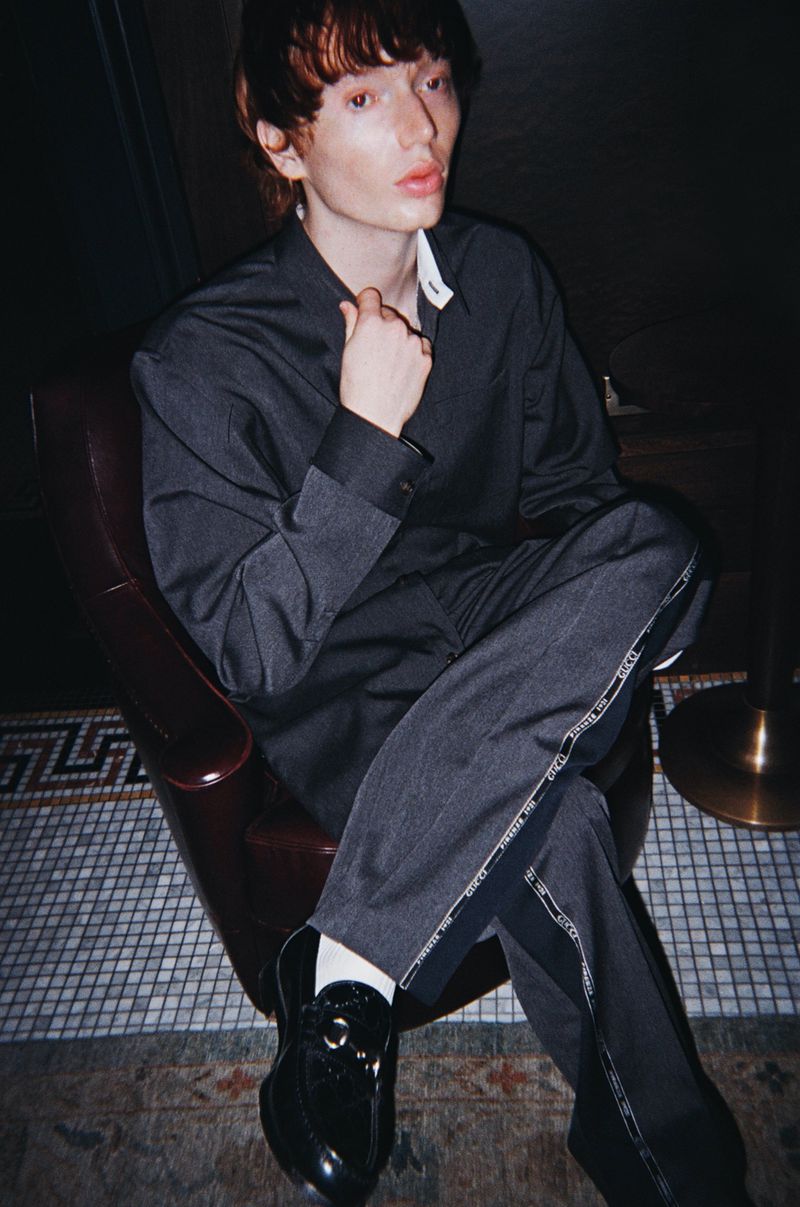 A young adult male sits in a red chair wearing a grey suit and black loafers, with a dark and moody atmosphere, indoors.