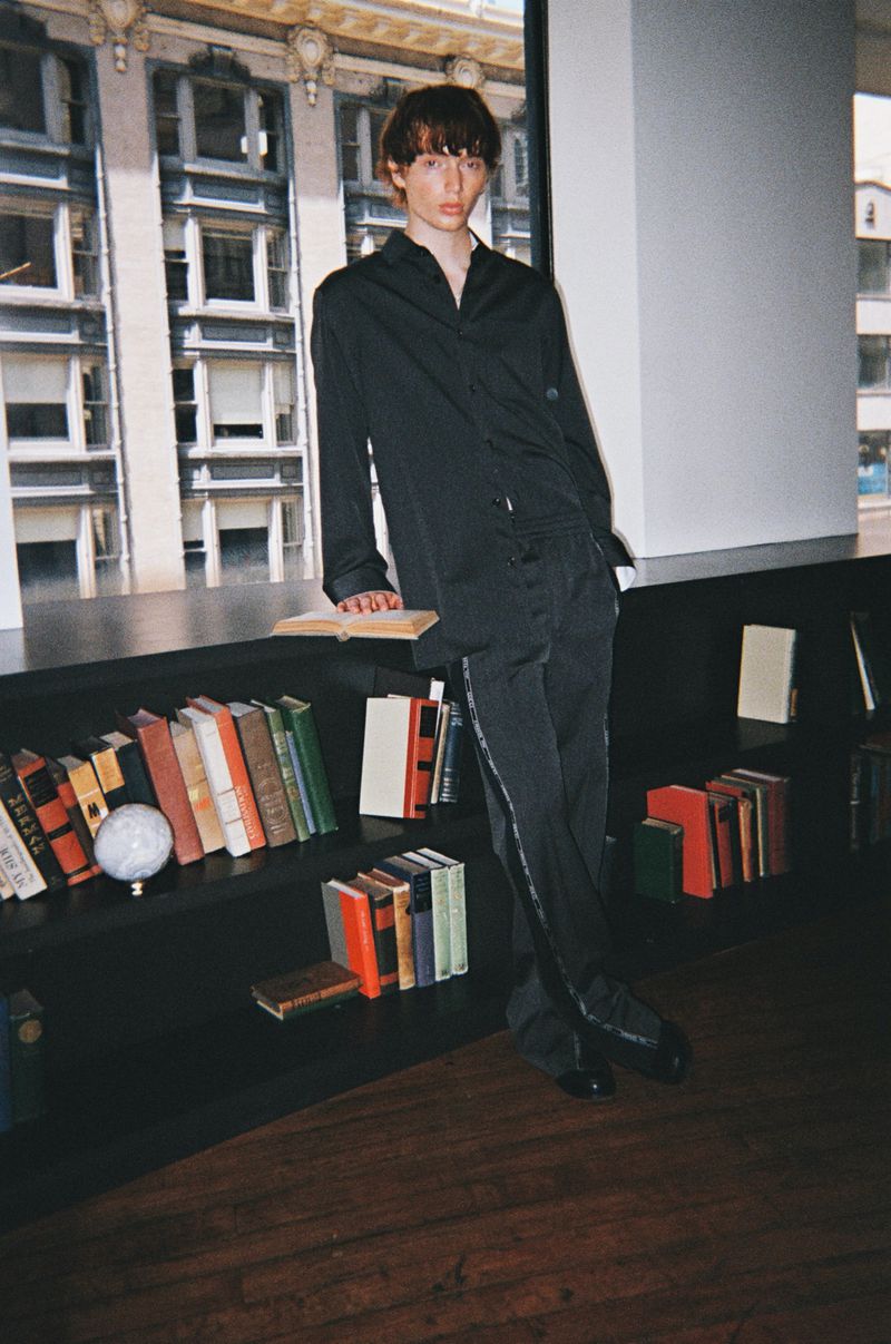 Young male model wearing a dark shirt and trousers leaning against a dark shelf filled with books in front of large windows.