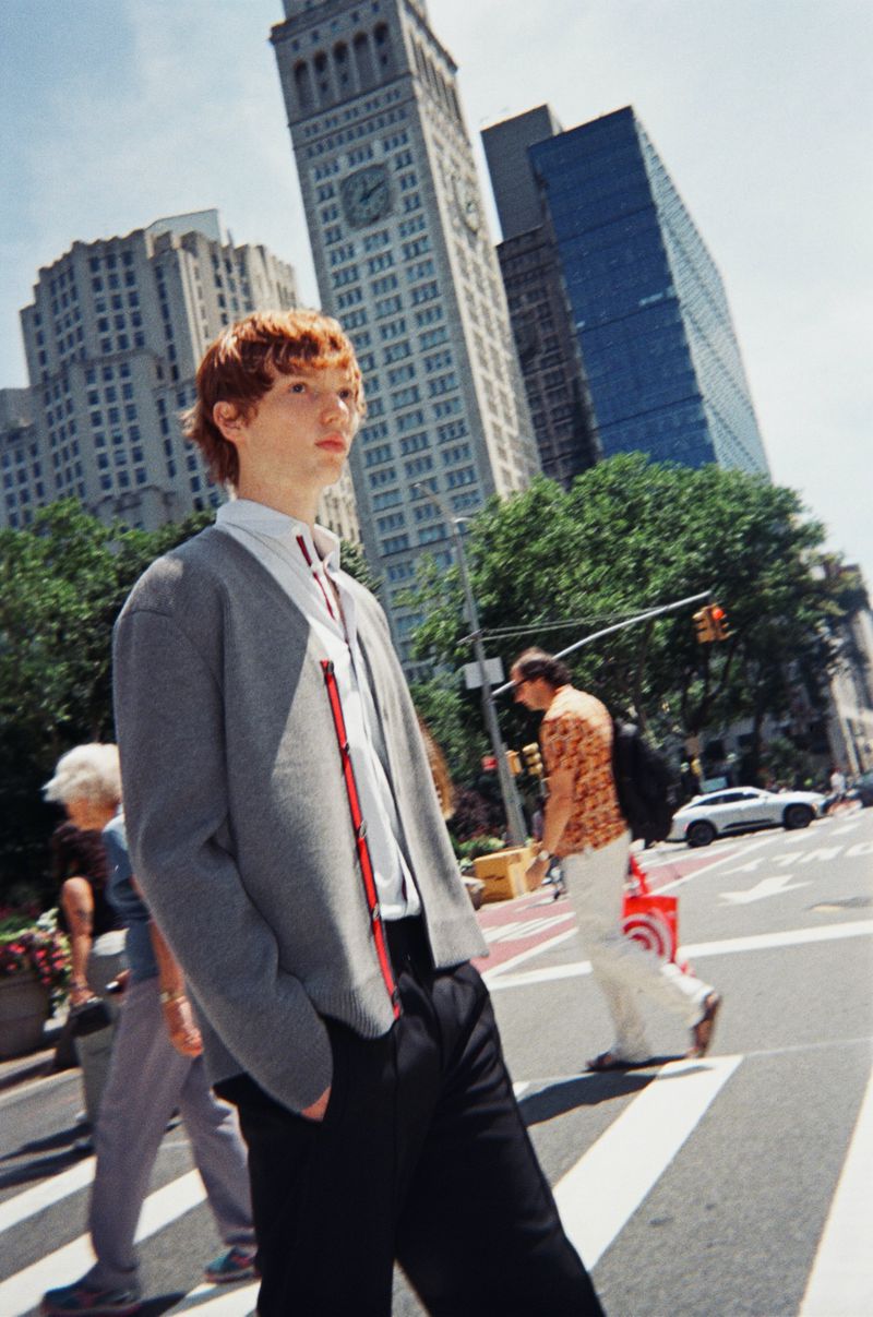 Young redhead man in grey cardigan walks across a New York City crosswalk with tall buildings in the background on a bright day.