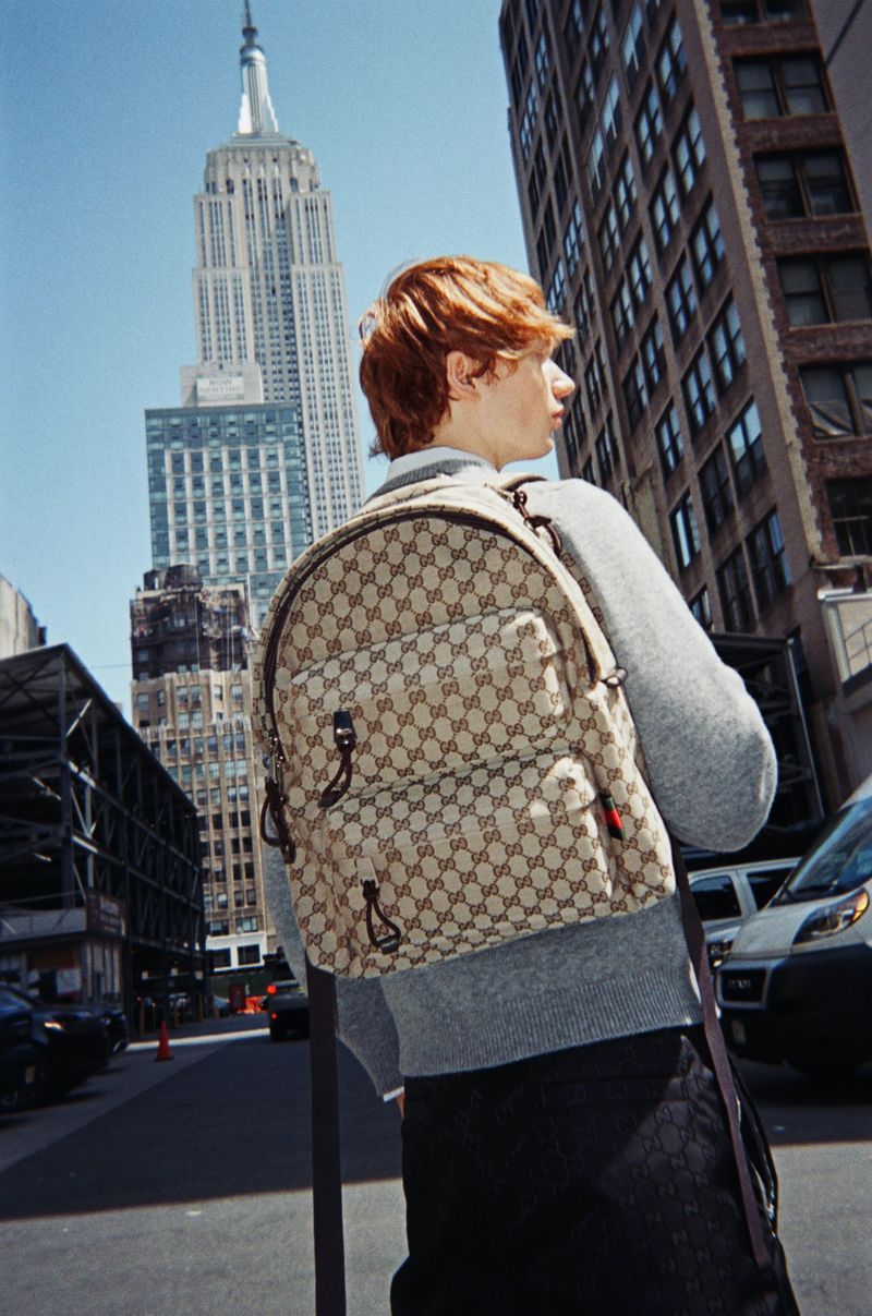 A person wearing a Gucci backpack stands on a city street with high-rise buildings, including the Empire State Building, on a sunny day.