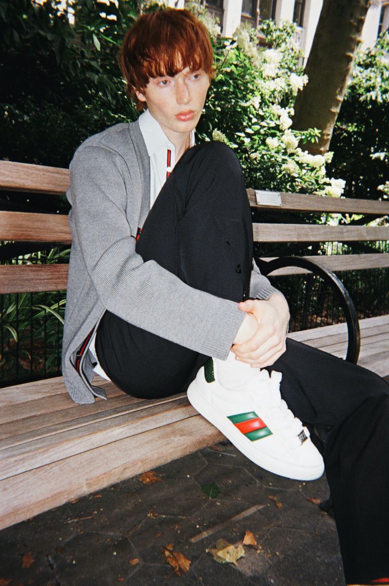 Young adult with auburn hair wearing a gray cardigan, black pants, and designer sneakers sitting on a park bench outdoors.