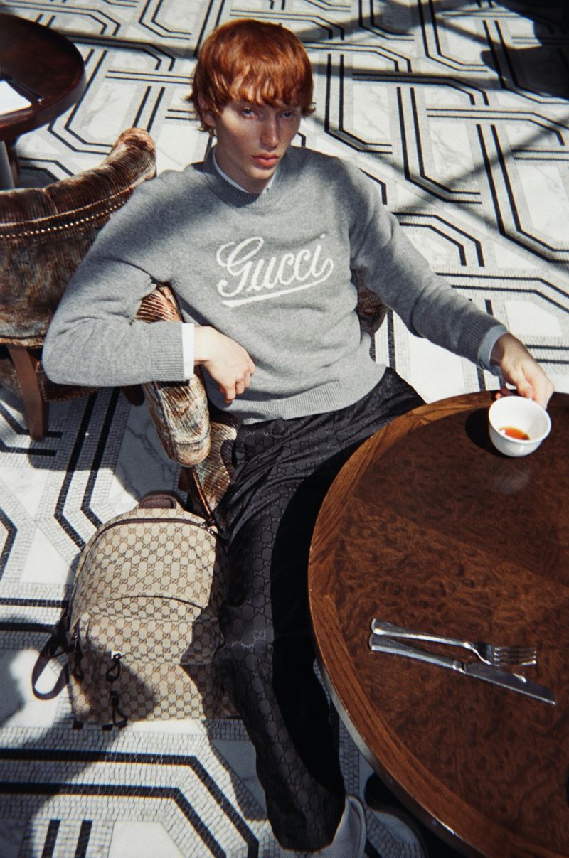 Red-haired male model wearing a grey Gucci sweater seated at a round wooden table in a cafe